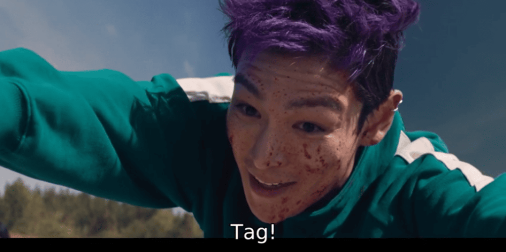 Squid Game 2: A person with purple hair in a green tracksuit with their arms outstretched