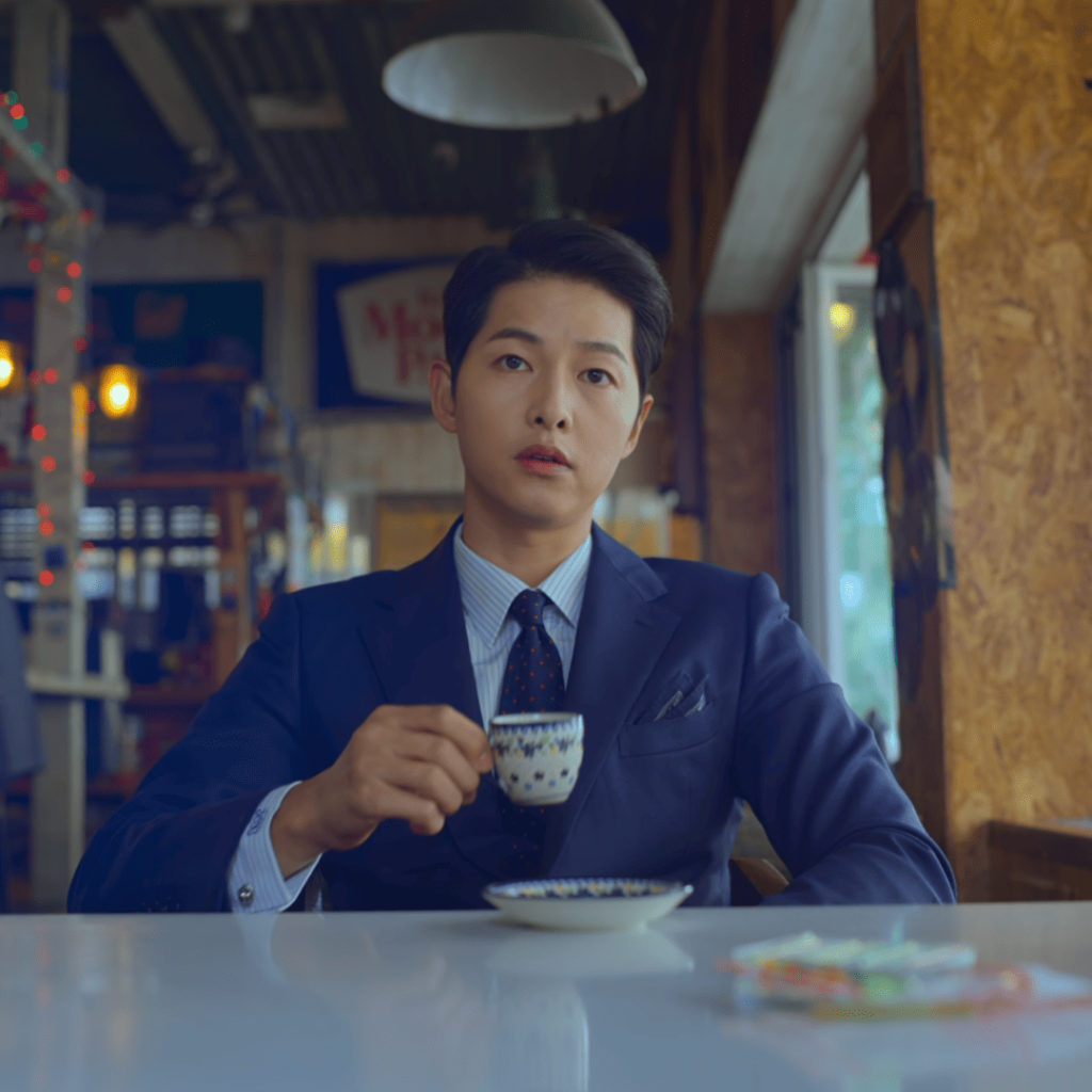 revenge k-dramas: A person in a suit and tie sitting at a table with a cup of coffee.