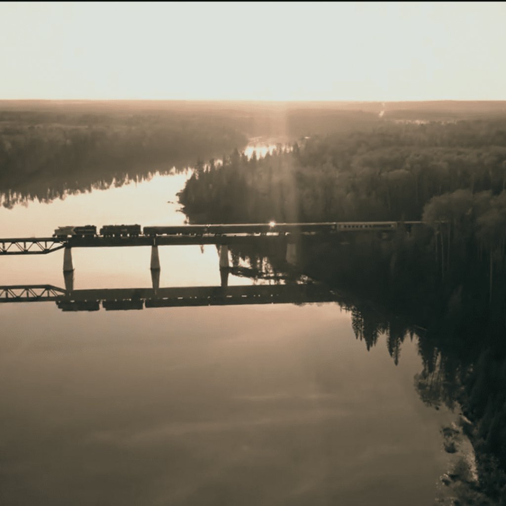 A train crosses a bridge over a still river surrounded by dense forest, bathed in soft golden light in Handmaid’s Tale Season 6
