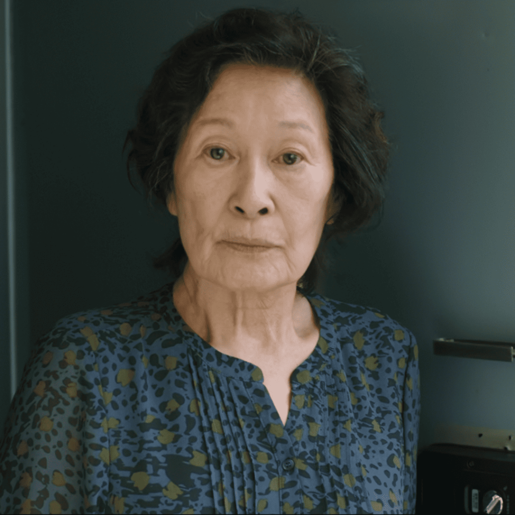 Heavenly Ever After:  Elderly Korean woman standing in front of a door, wearing a blue patterned blouse and looking directly at the camera with a calm, serious expression