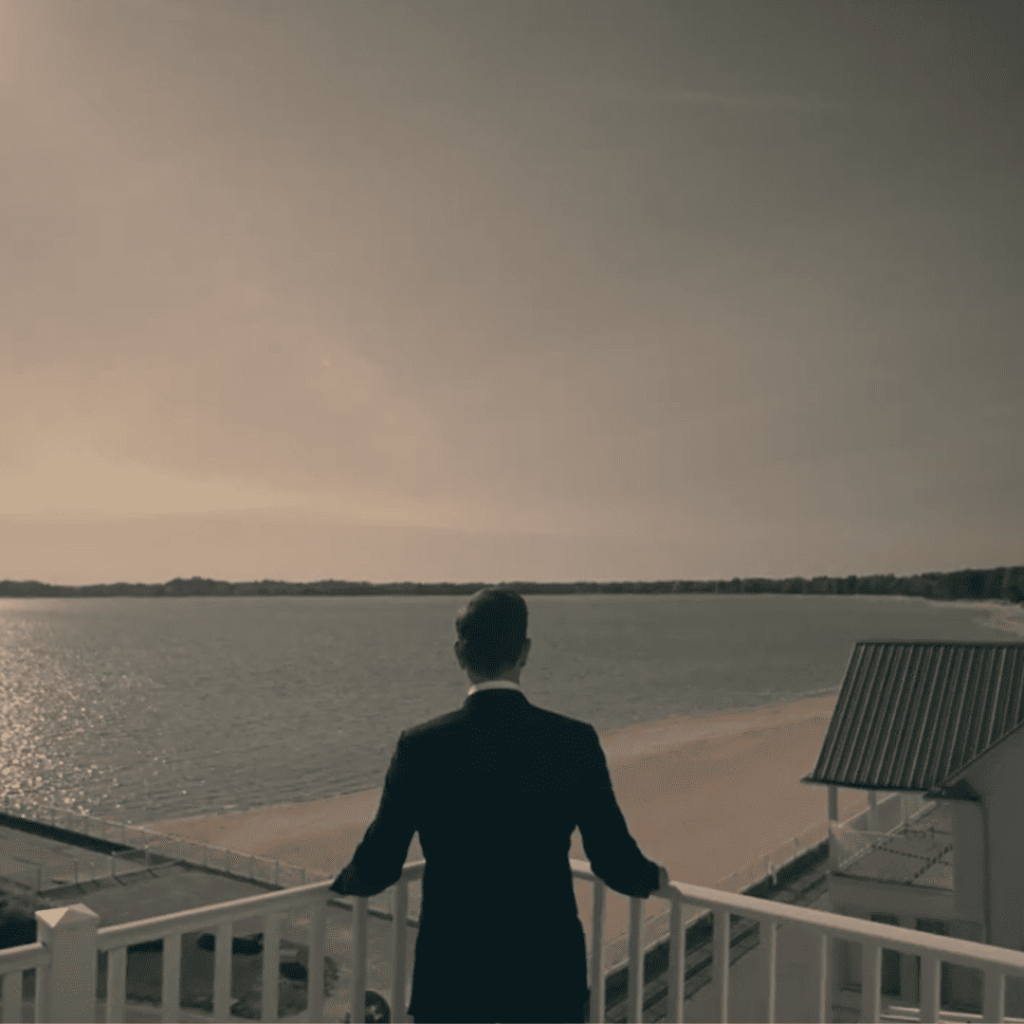 A high-ranking commander in a dark suit gazes out over a quiet, sunlit shoreline in Handmaid's Tale Season 6