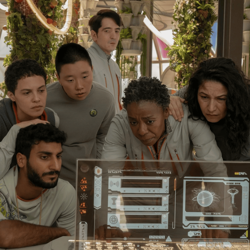 Murderbot Show Explained: The human crew crowds around a futuristic screen, cluelessly trying to solve problems while Murderbot tunes out.