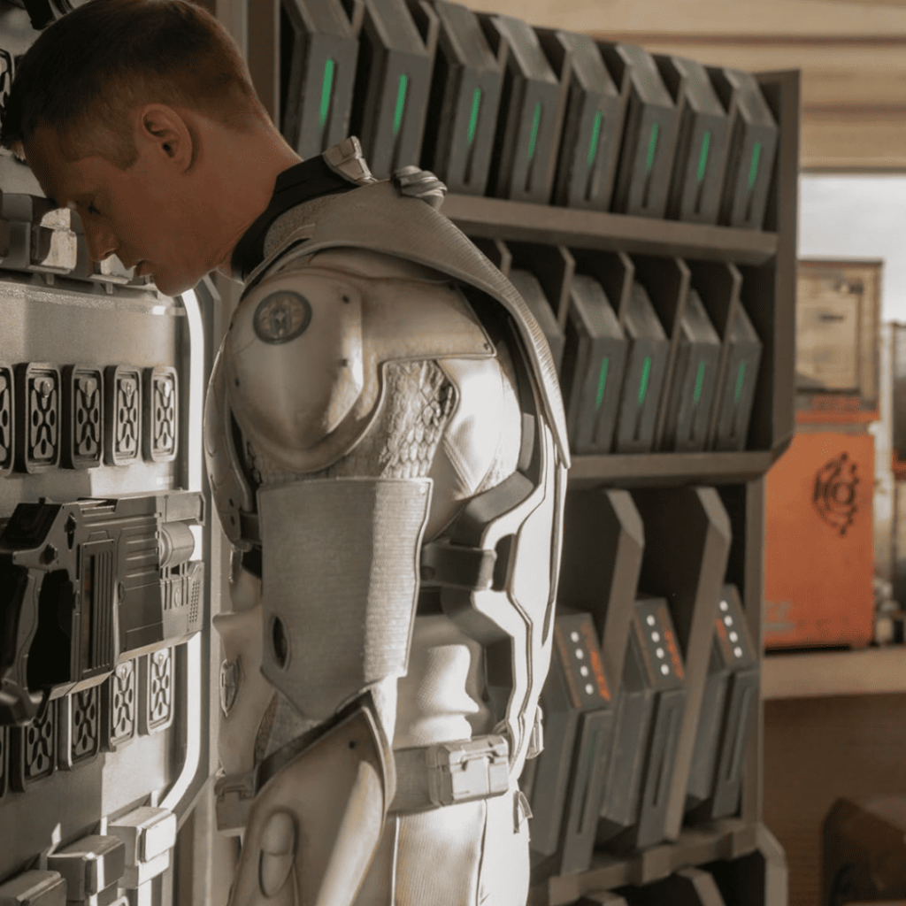 Murderbot Show Explained: A weary SecUnit in white armor rests its head against a wall of weapons and storage units, symbolizing exhaustion and reluctant heroism.