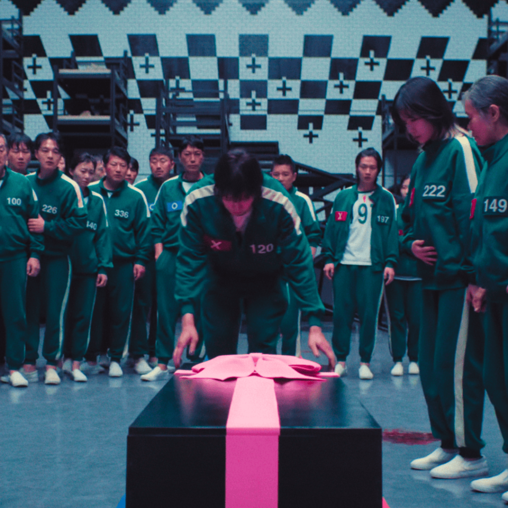 Squid Game contestants in green tracksuits gather solemnly around a black coffin wrapped with a pink ribbon.
