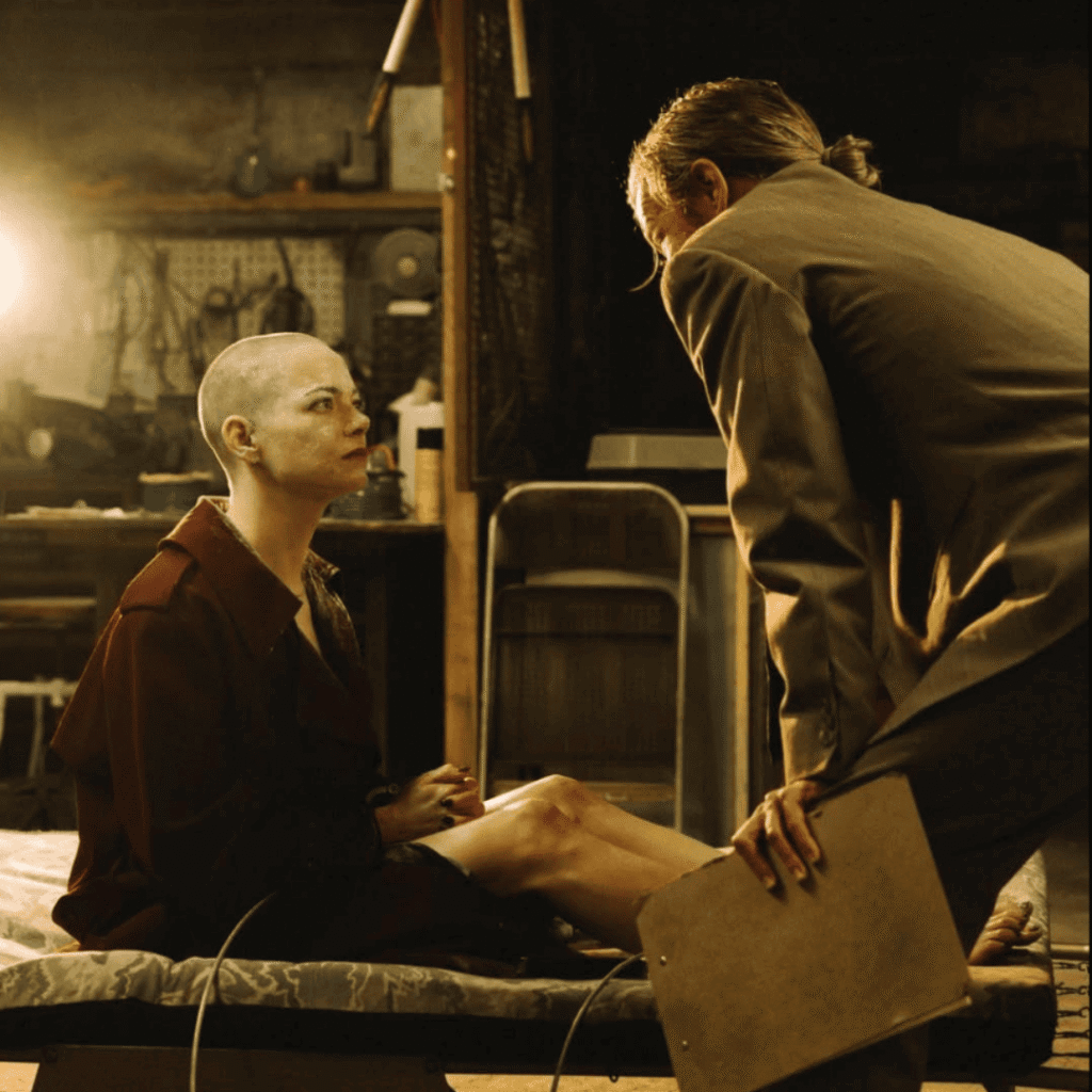 Bugonia Review: Teddy and Michelle facing each other in the basement, she is on a makeshift bed while he holds a clipboard and interrogates her.