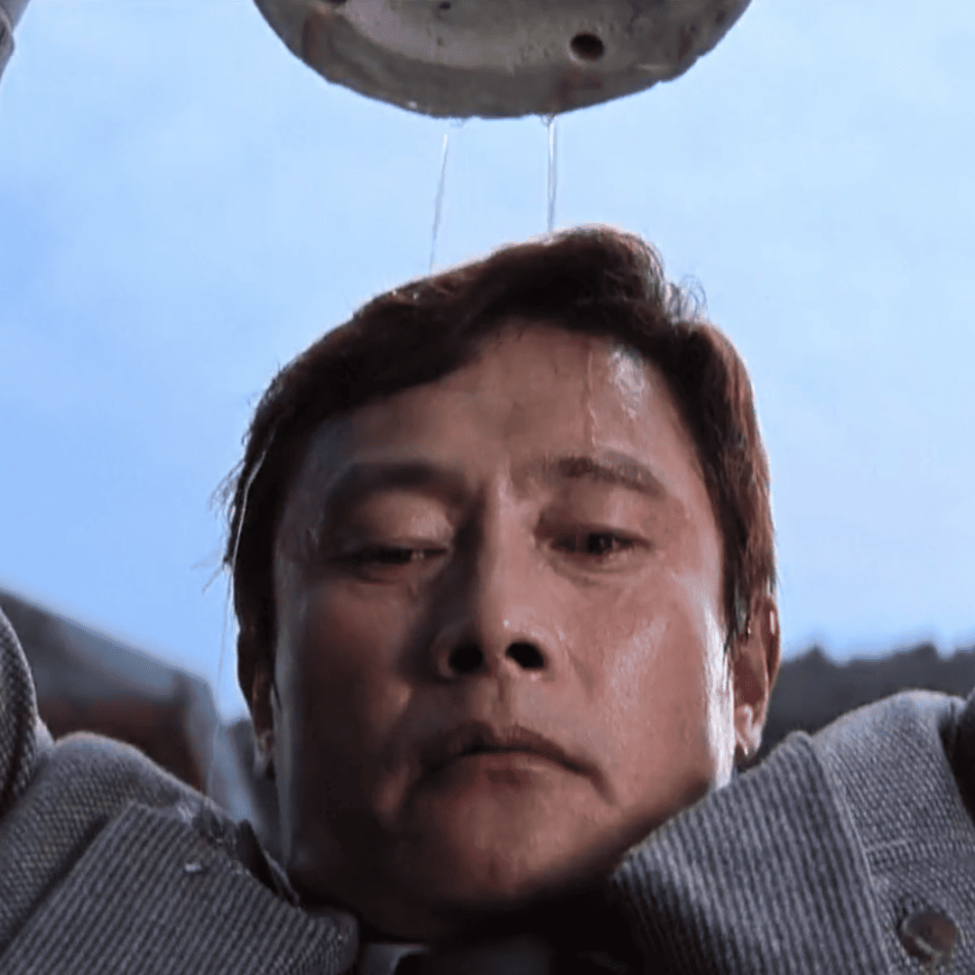 : A close up of Man-su on a rooftop for this No Other Choice Review holding a heavy vase above Choi Seon-chul.