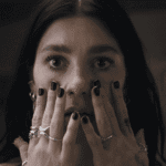 A cinematic still of Rachel from the Something Very Bad Is Going to Happen review, her hands covered in black nail polish and silver rings pressed against her face in a look of total disbelief.