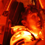 A harrowing close-up for the Project Hail Mary Review showing an injured Ryland Grace, with a busted nose and covered in a red light filter, reaching for a critical button inside the ship.
