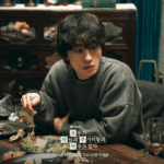 A production still for the We Are All Trying Here review released by JTBC, featuring Koo Kyo-hwan as Hwang Dong-man sitting at a restaurant table with a plate of food in front of him.
