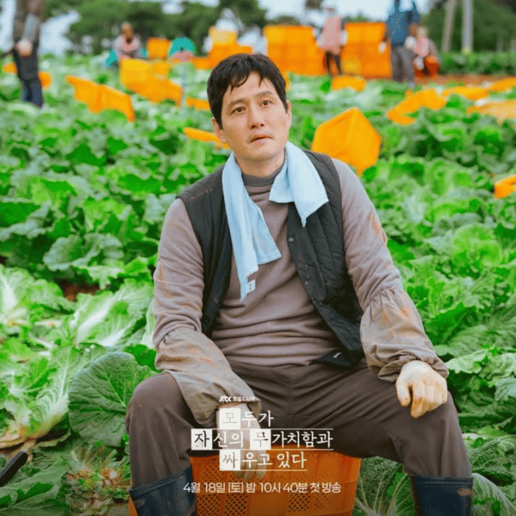 A production still for the We Are All Trying Here review released by JTBC, epicting Park Hae-joon as the former poet Hwang Jin-man sitting isolated on a plastic basket in the middle of a vast green Napa cabbage field.
