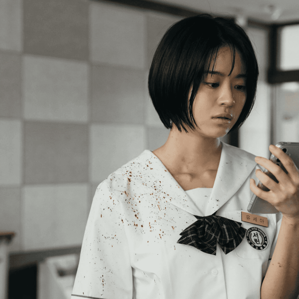 A visceral image for the If Wishes Could Kill Review of Se-ah in a blood-sprayed school uniform, focused intensely on her mobile phone screen.
