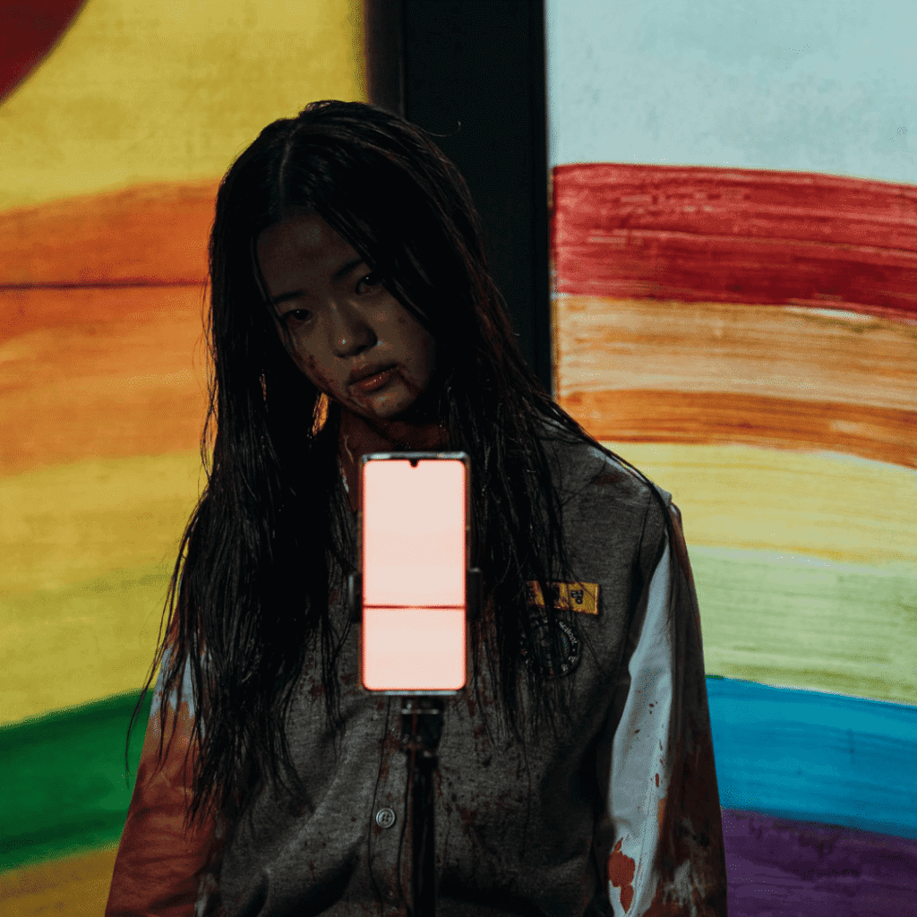 : A cinematic still for the If Wishes Could Kill Review of Hae-ryeong in the warehouse, positioned in front of a tripod with the iconic rainbow window in the background.