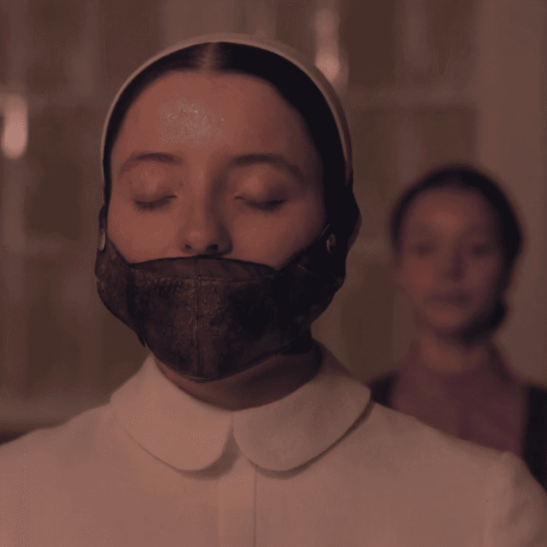 A close-up still image for The Testaments Review showing Daisy with a mouth mask covering her face while being accused of blasphemy with Agnes blurred in the background.