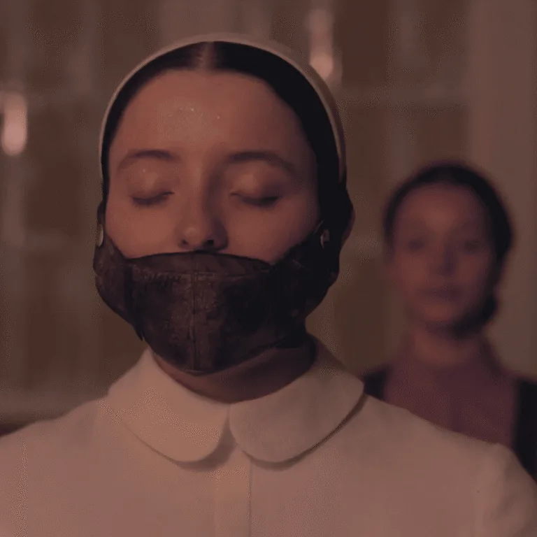 A close-up still image for The Testaments Review showing Daisy with a mouth mask covering her face while being accused of blasphemy with Agnes blurred in the background.