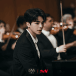 Alt Text: Official tvN still of Kang Pi-o sitting at the piano as an orchestra prepares for a competition in Four Hands, Two Sonatas.