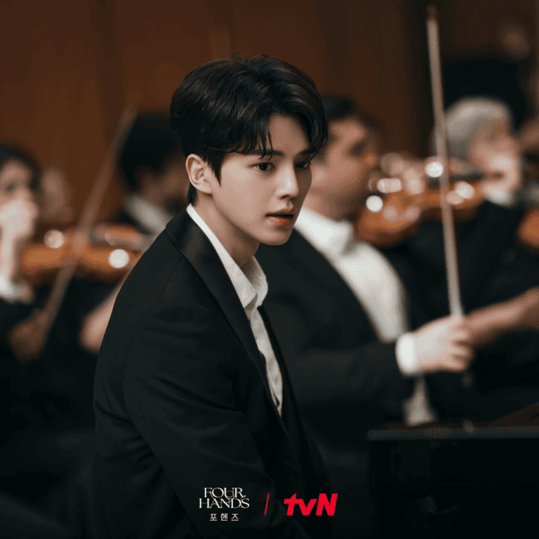 Alt Text: Official tvN still of Kang Pi-o sitting at the piano as an orchestra prepares for a competition in Four Hands, Two Sonatas.