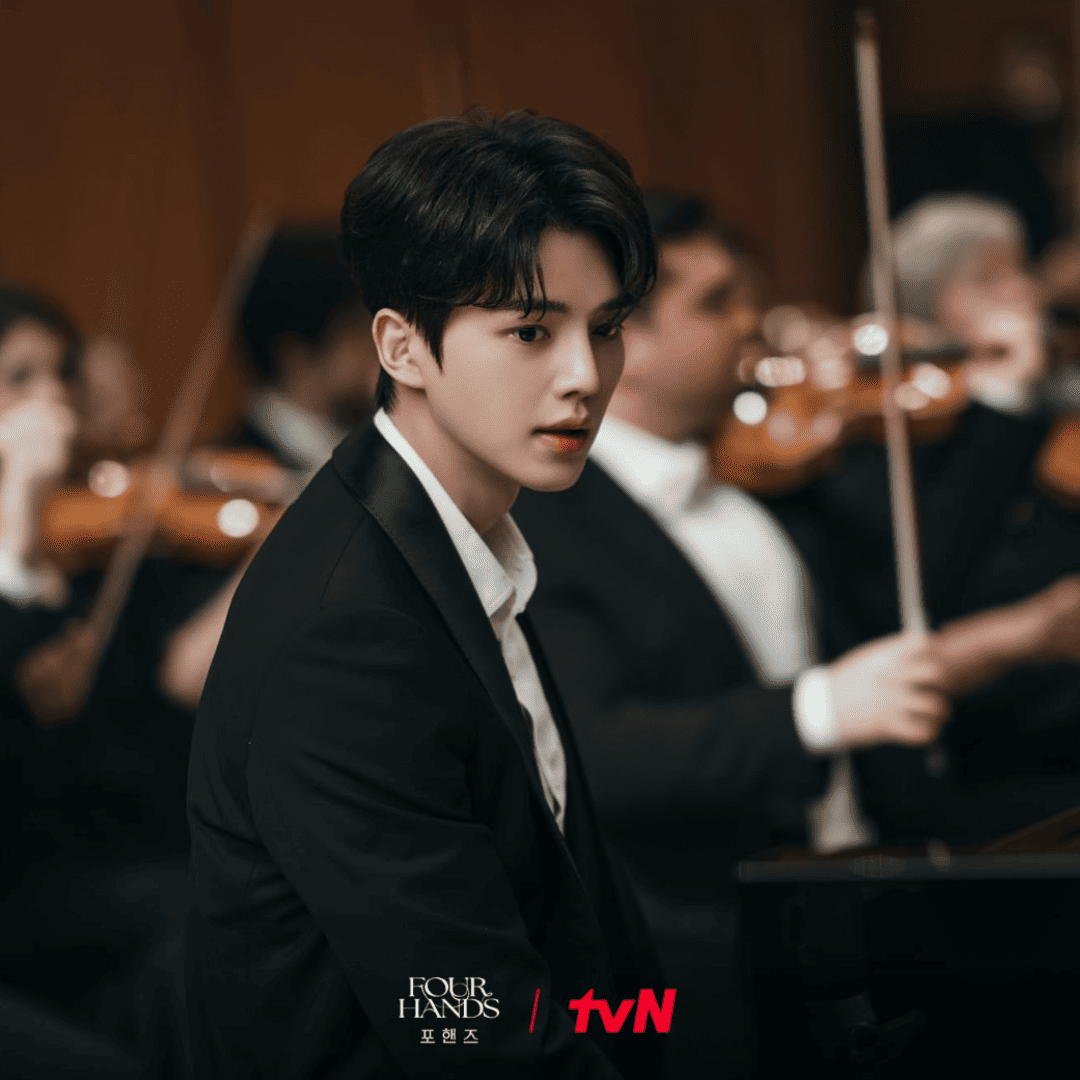 Alt Text: Official tvN still of Kang Pi-o sitting at the piano as an orchestra prepares for a competition in Four Hands, Two Sonatas.
