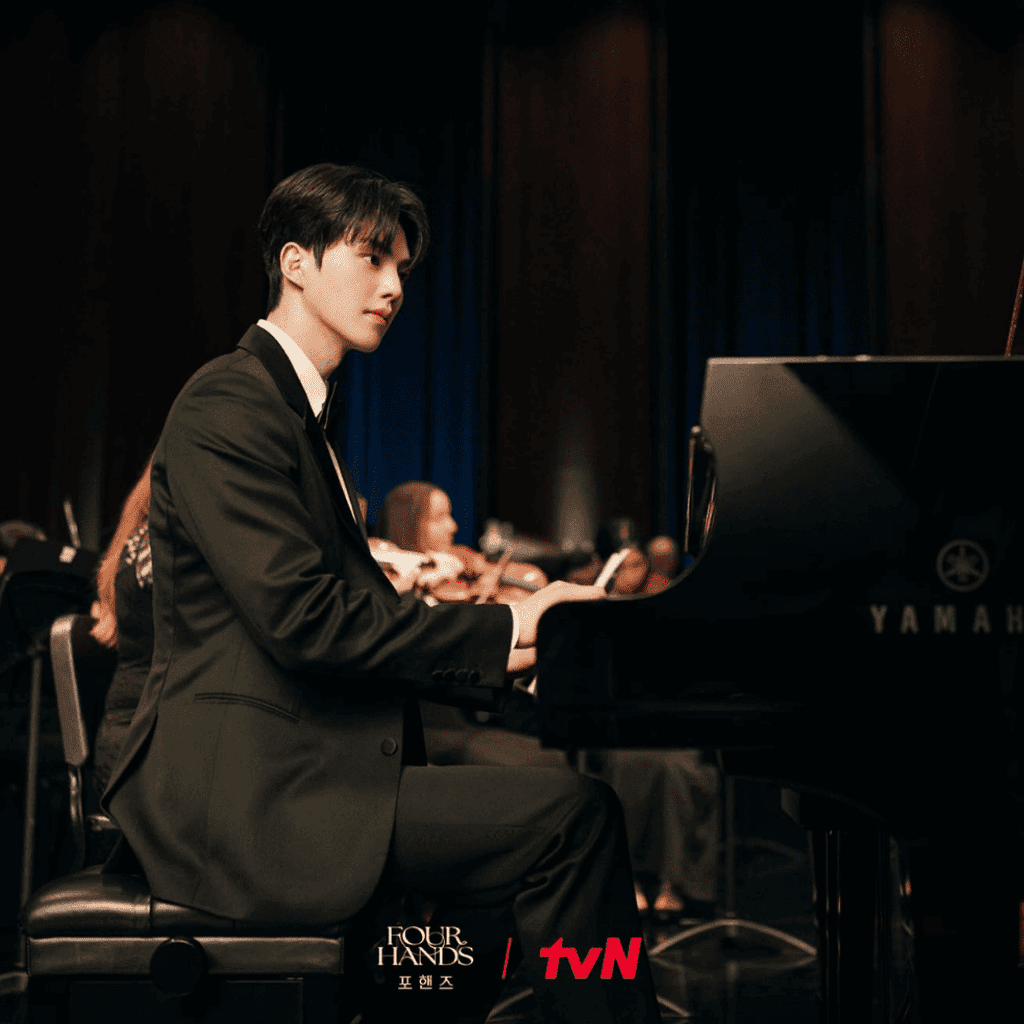 Alt Text:
Close-up profile of Kang Pi-o sitting at a Yamaha piano with the orchestra blurred behind him in Four Hands, Two Sonatas.