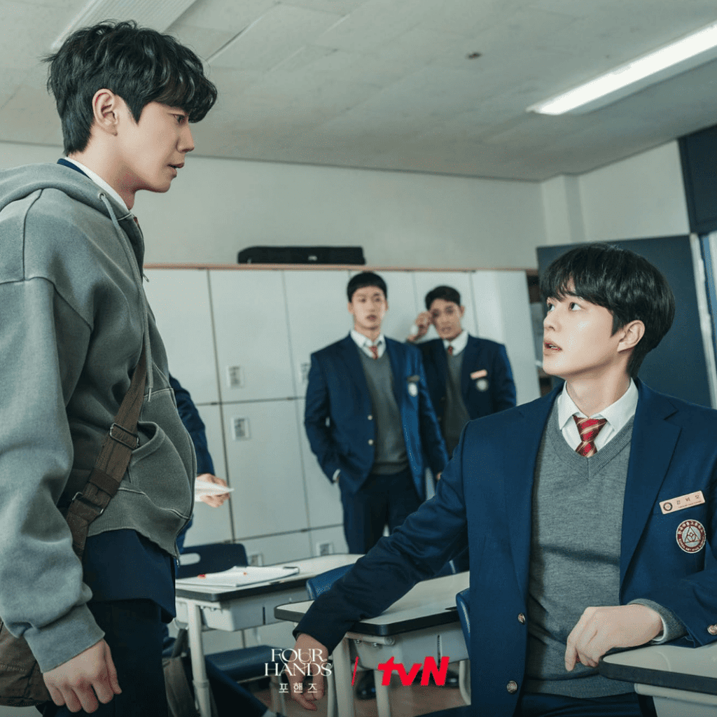 Alt Text:
Kang Pi-o sits facing the standing Choi Jeong-yo during a tense classroom confrontation, with two students in the background in Four Hands, Two Sonatas.